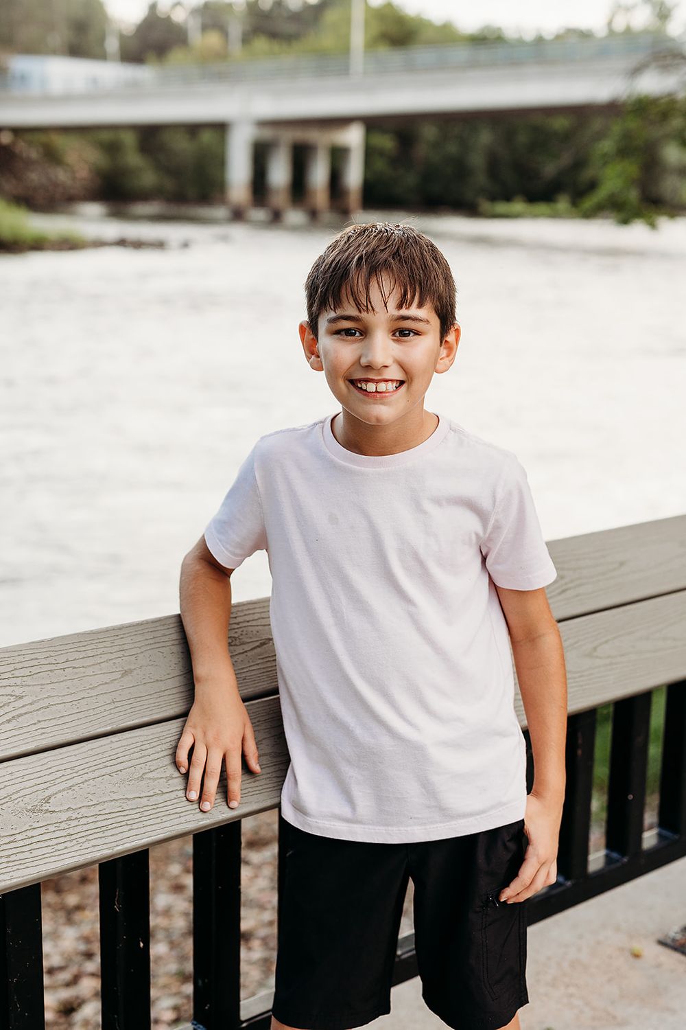 A young child wearing a white t-shirt and dark shorts stands smiling while leaning against a wooden railing outdoors.