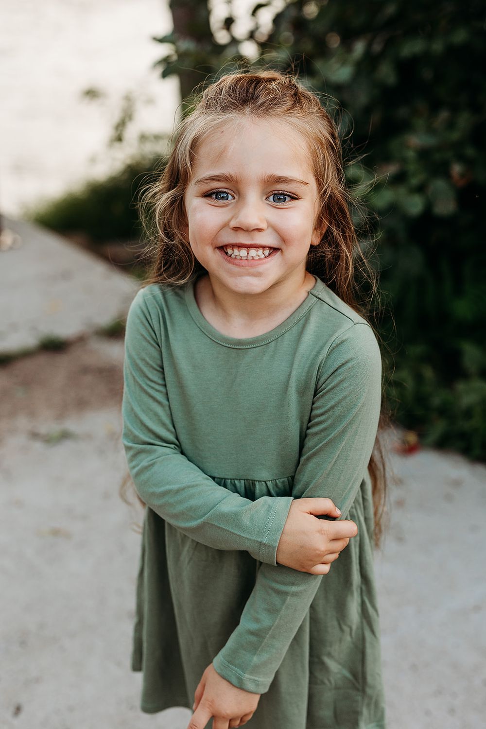 Young child wearing a long-sleeved green dress smiles brightly outdoors against a natural background.