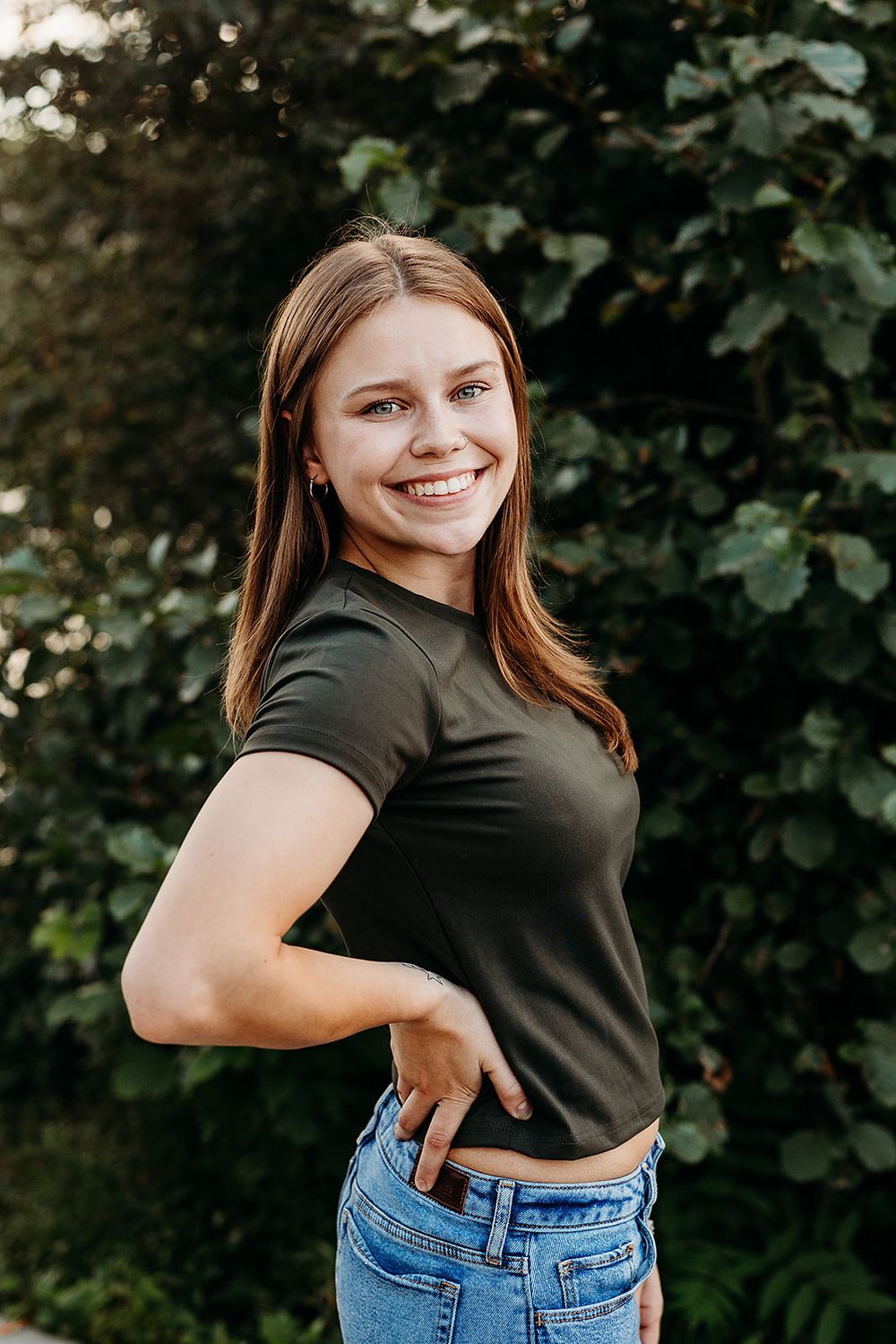 Person in black t-shirt and blue jeans poses against green foliage background in outdoor portrait.