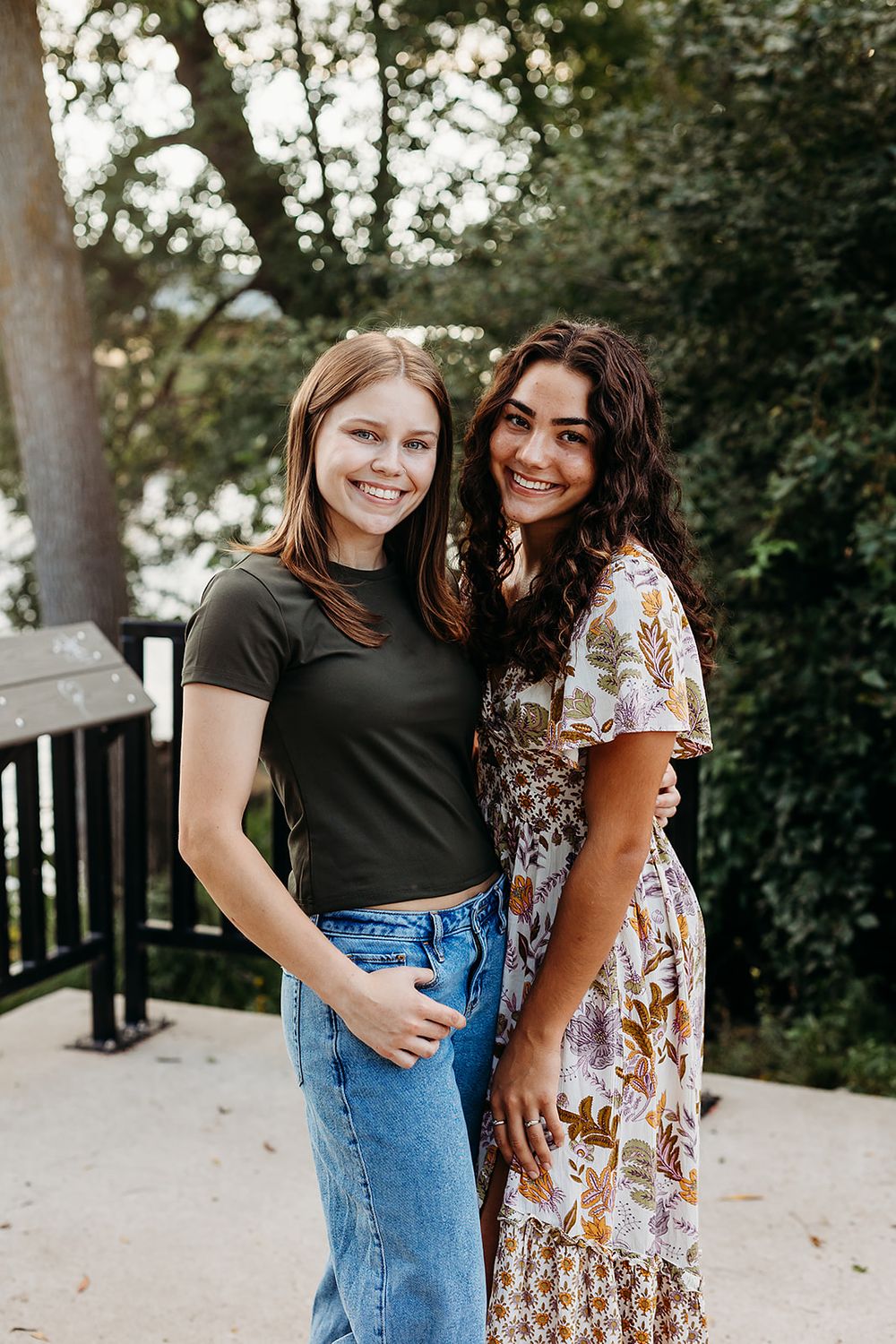 Three friends smiling and posing together outdoors in front of trees wearing casual attire.