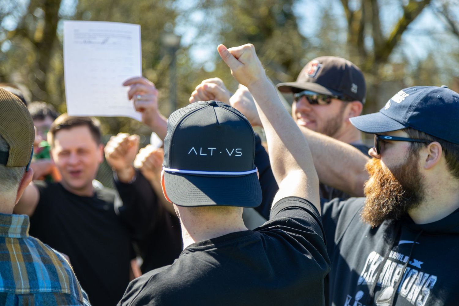 Group of men outdoors at a rally, one wearing an ALT VS cap, others raising fists in background.