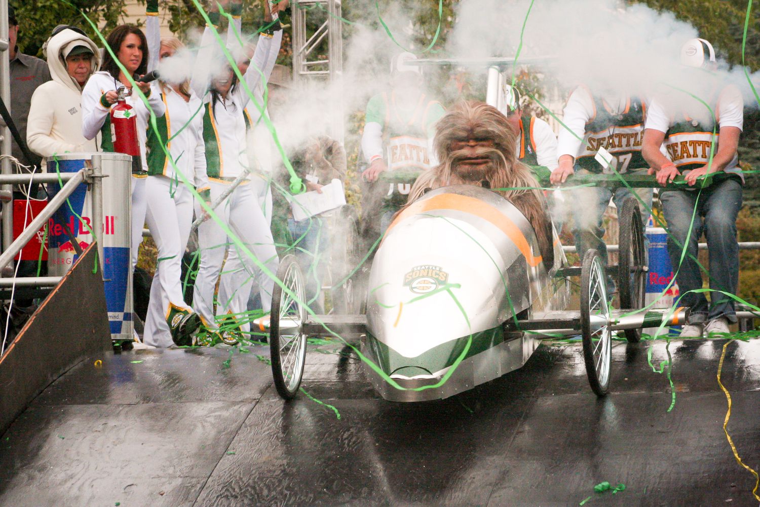 A soap box derby racer speeds down a track surrounded by green smoke and cheering spectators at an event.