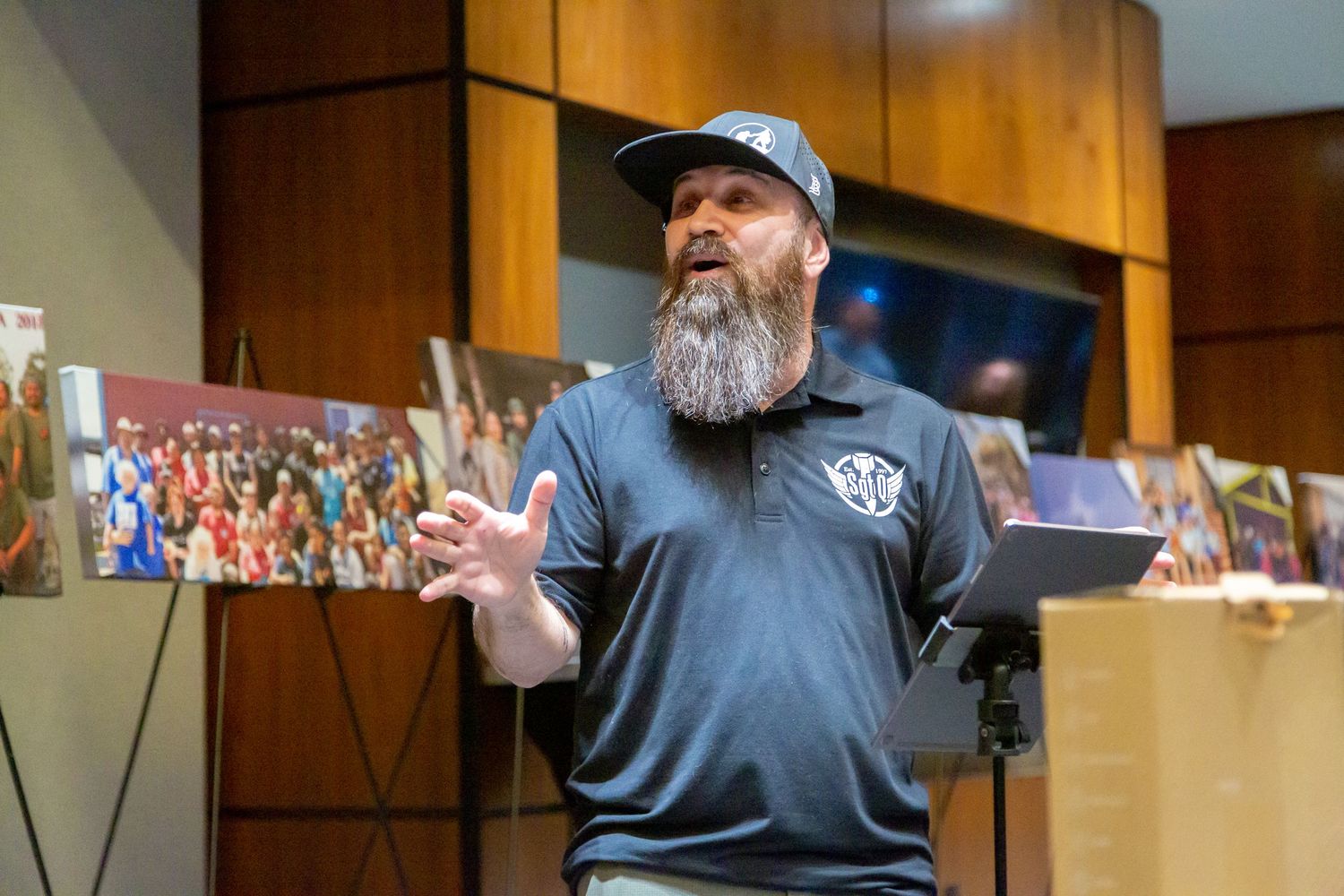A casually dressed speaker in a baseball cap gestures while giving a presentation in front of a display wall.