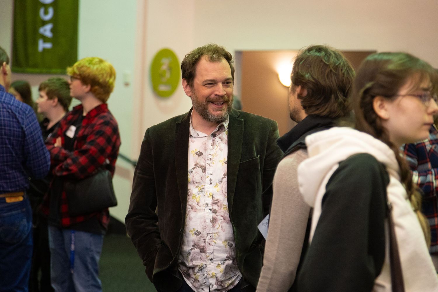 Man in floral shirt smiling at an indoor networking event with people gathered in background.
