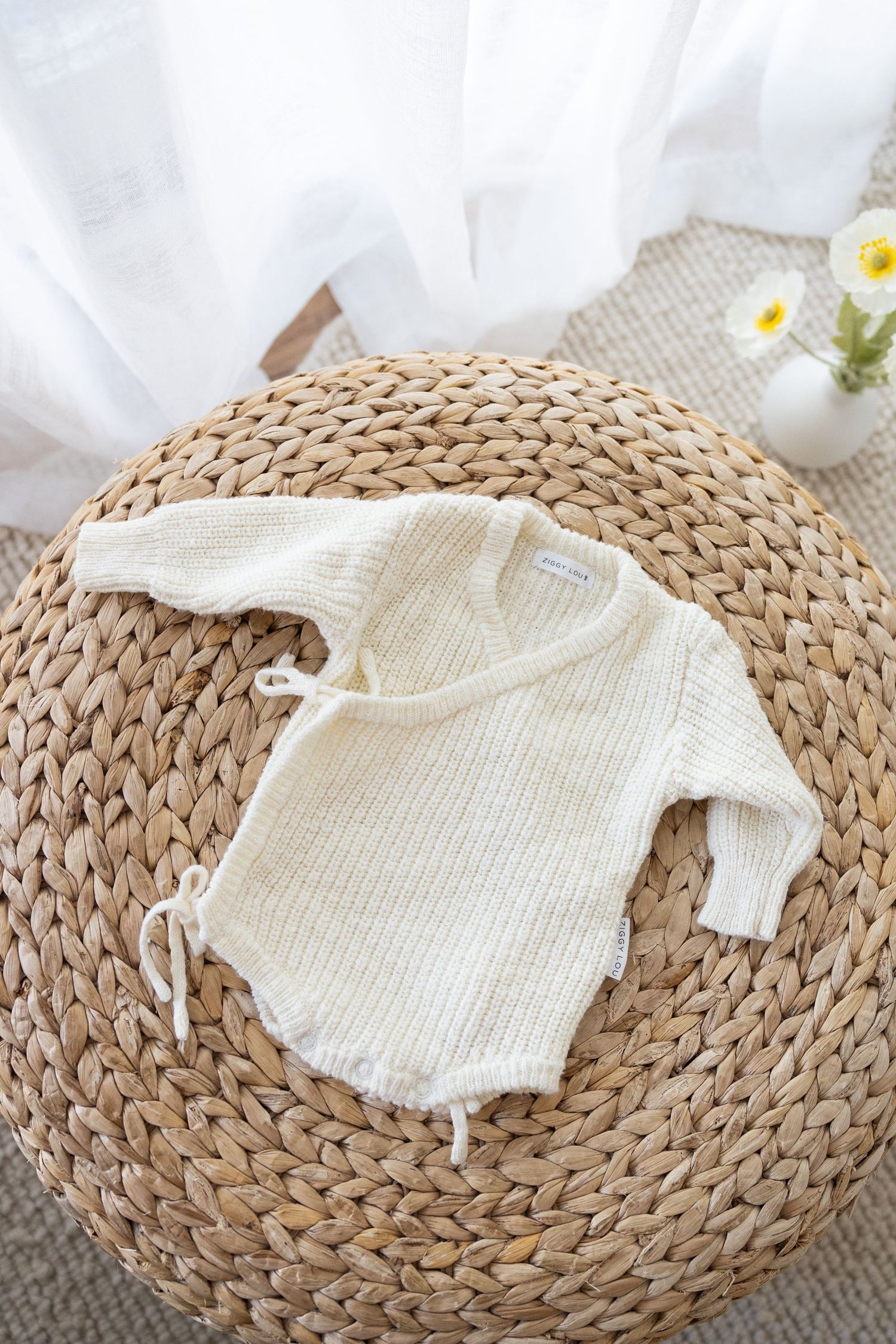 White knit baby romper displayed on round woven straw mat with daisies in vase on rustic background.