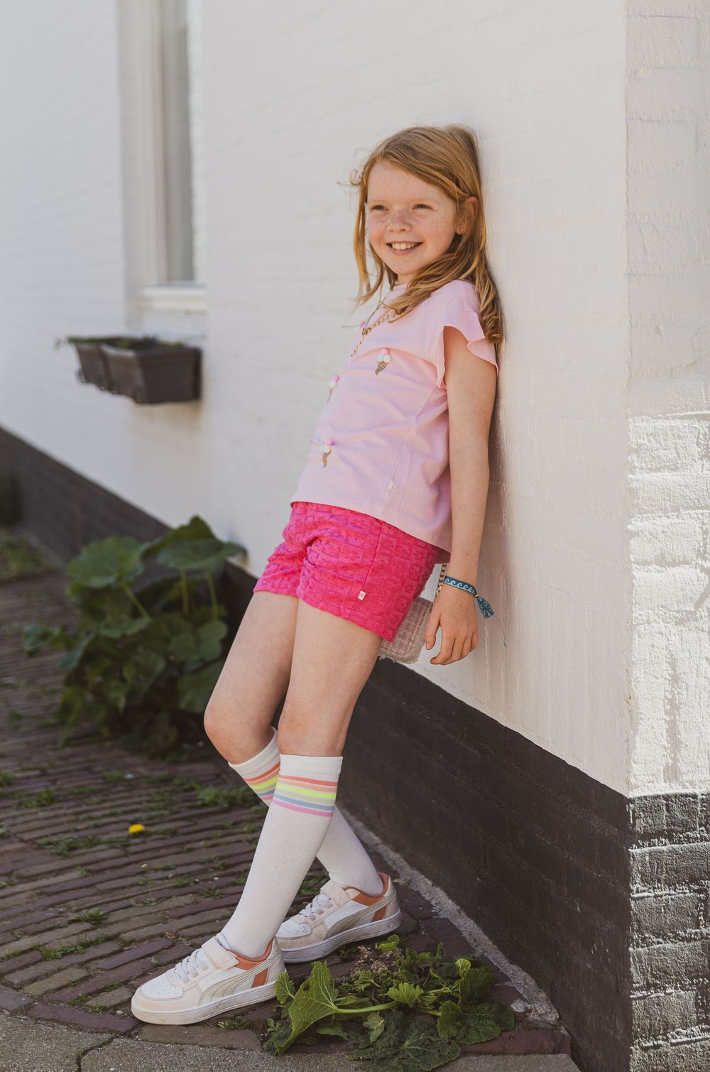 young child in a pink top and bright pink shorts leans against a white wall wearing knee socks and white sneakers. Kinderkledingwinkel foto