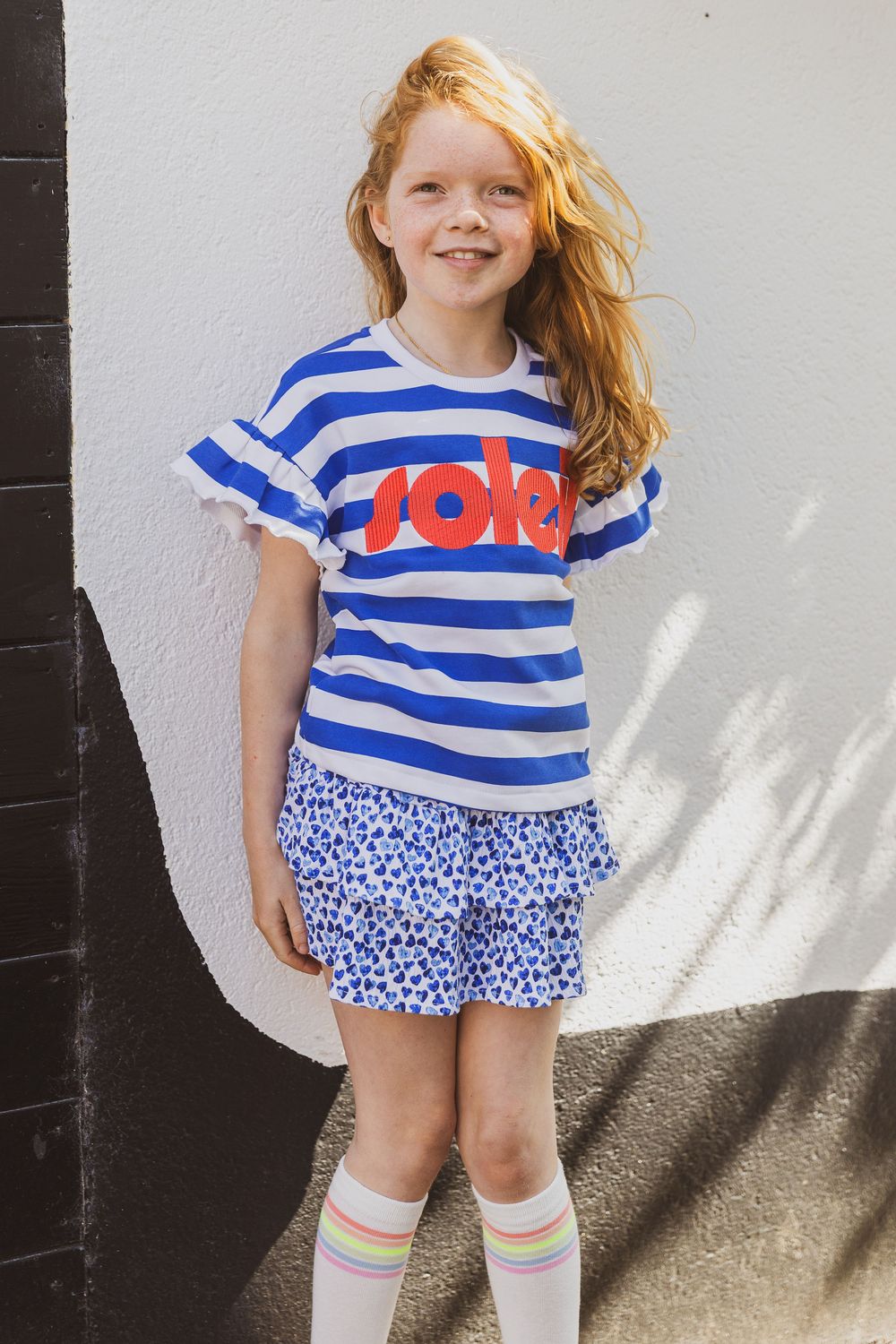 A child in a blue and white striped t-shirt with the word 'love' and a floral skirt poses against a white wall. Kinderportret foto