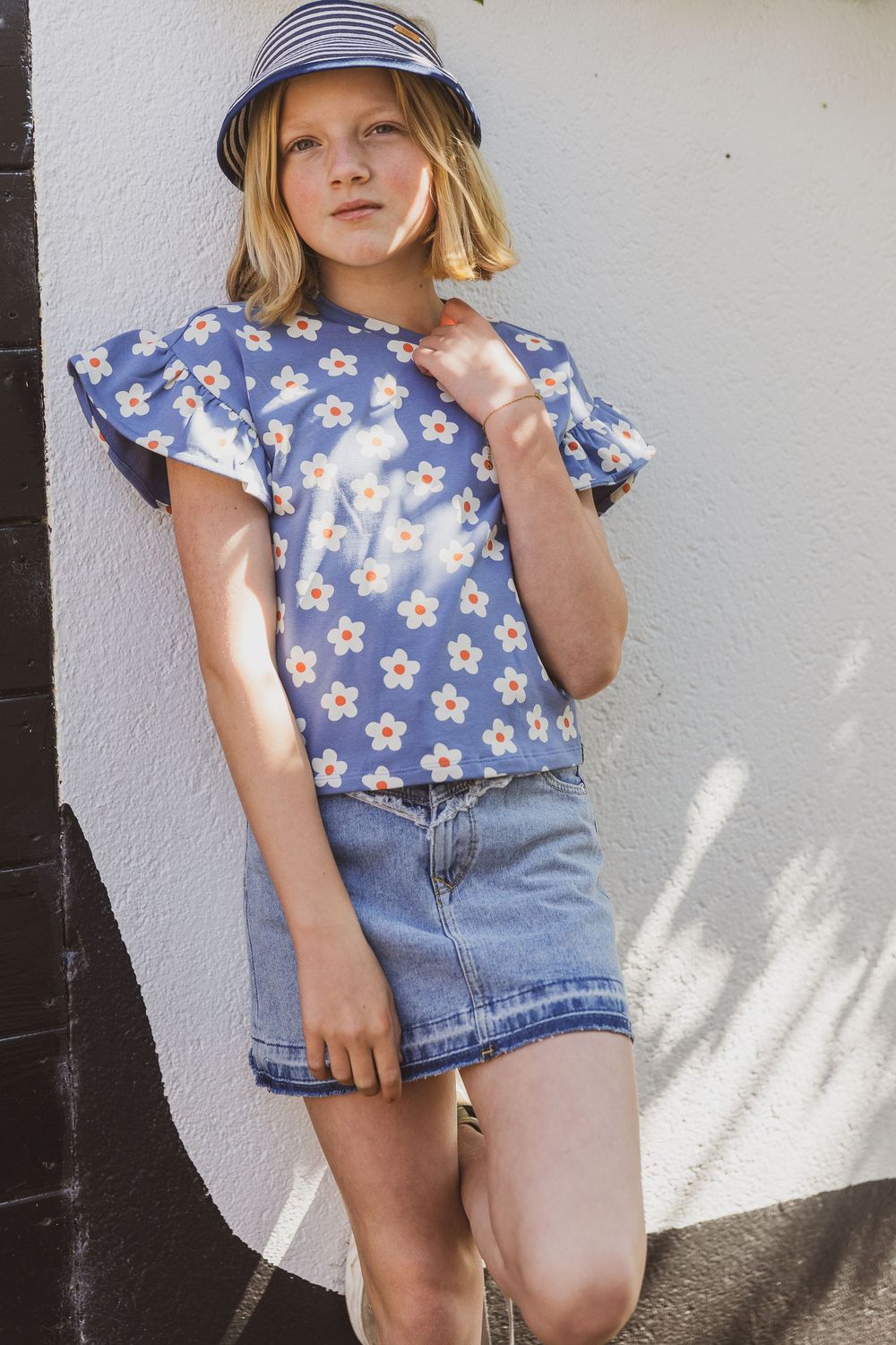 Child wearing blue floral ruffle sleeve top, striped hat and denim skirt poses against white wall in summer outfit. kinderkledingwinkel foto