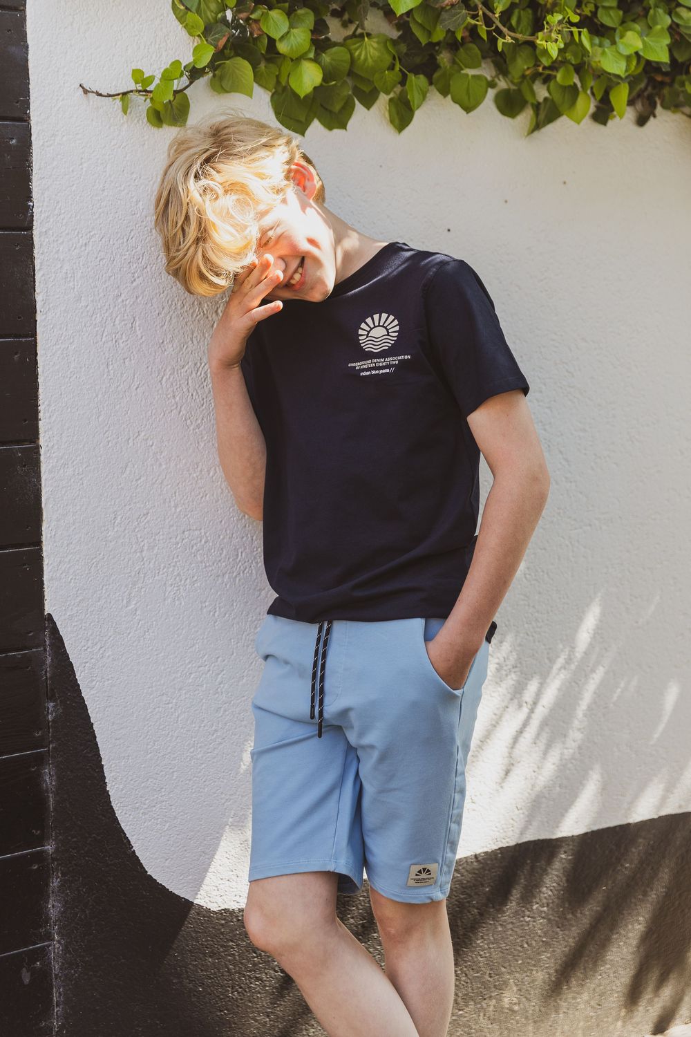 Child wearing black t-shirt and blue shorts poses casually against white wall with green vines overhead. Kinderportret