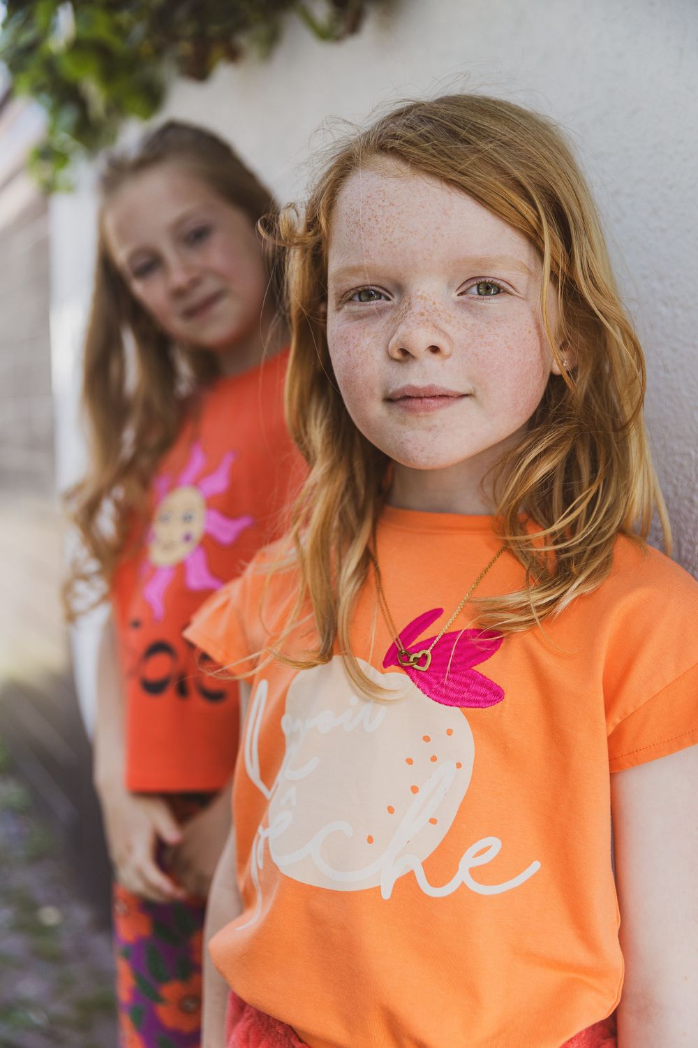 Two children in bright orange and coral t-shirts stand against a white wall outdoors. Editorial fotografie