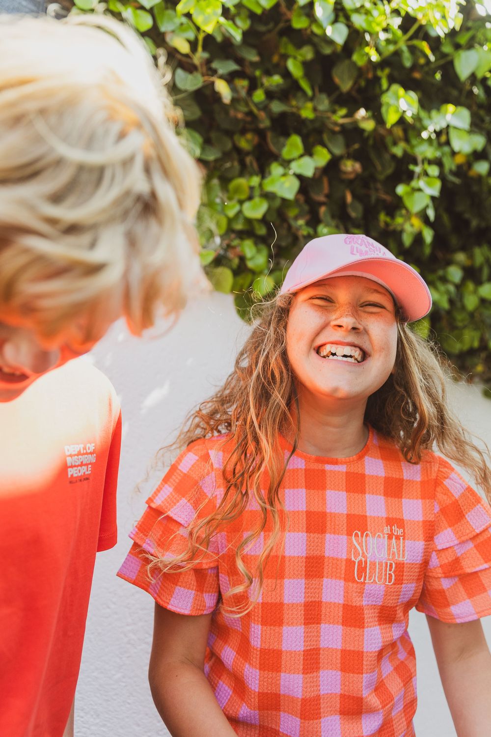 A person wearing a pink baseball cap and orange plaid shirt stands outdoors in front of green foliage laughing joyfully. Editorial foto