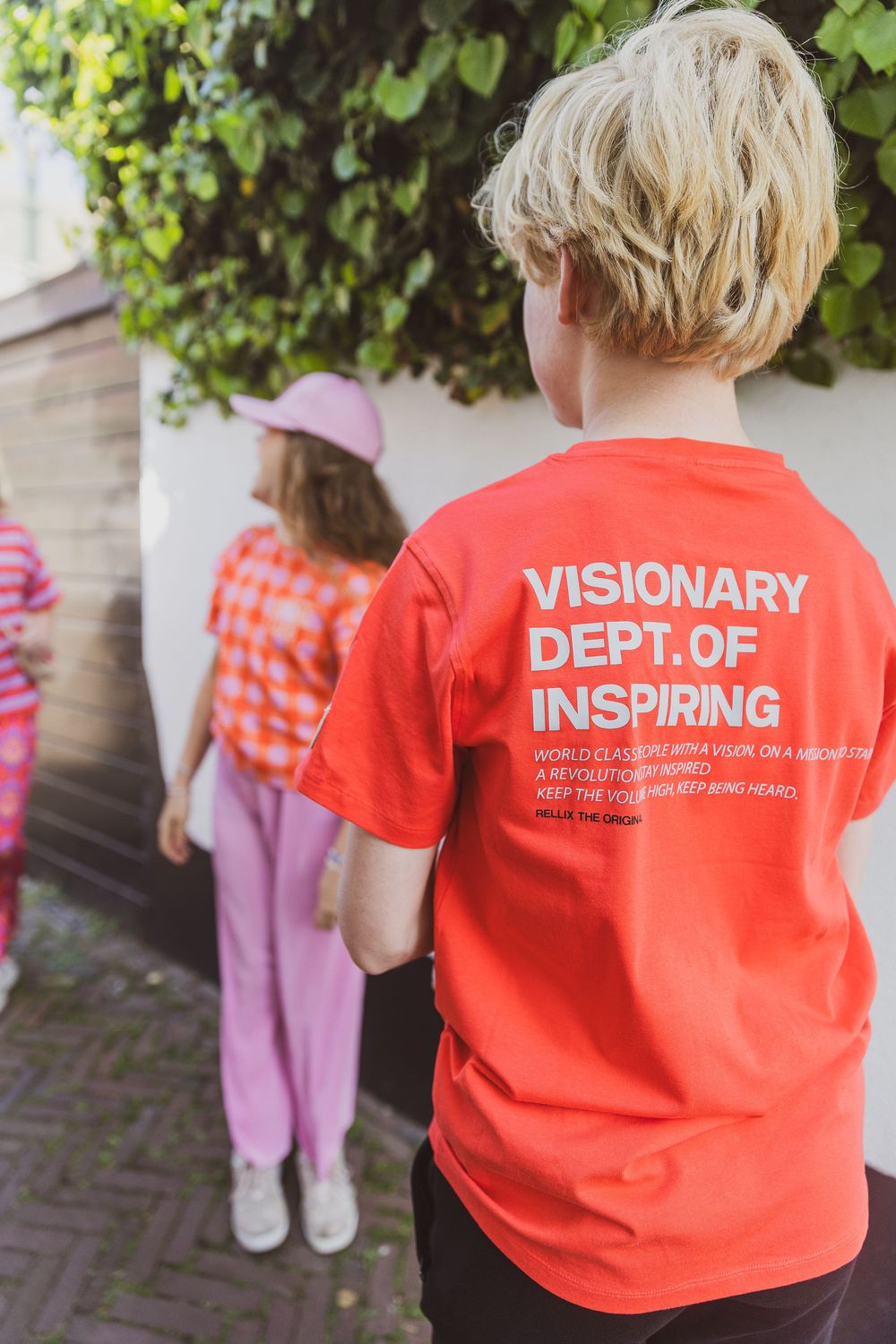 Person wearing a bright orange t-shirt with 'Visionary Dept of Inspiring' text standing outdoors near green foliage. editorial style foto