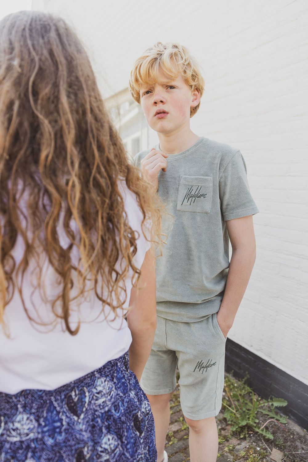 Young person wearing a grey t-shirt and matching shorts stands casually while conversing outdoors beside a wall.