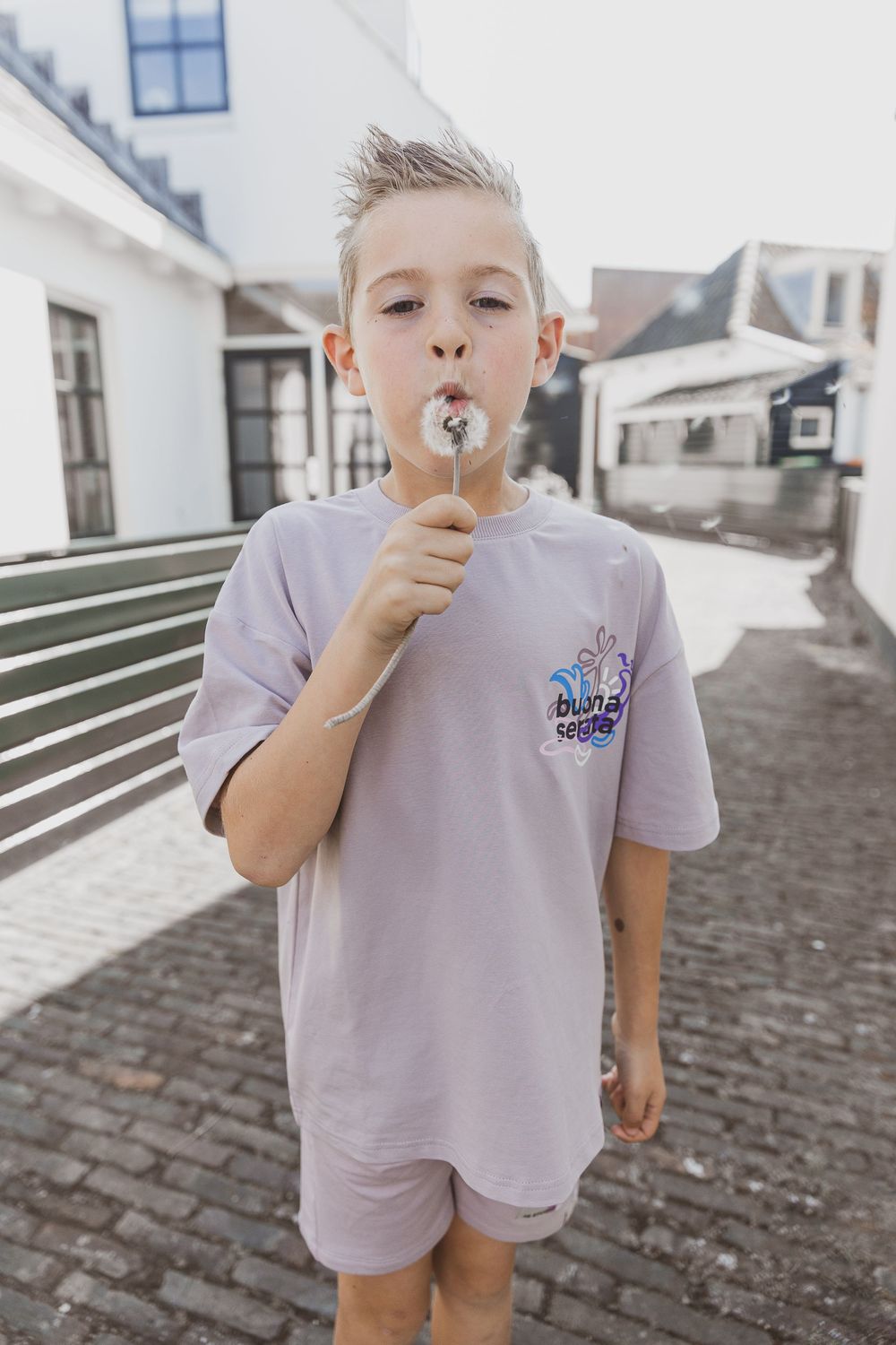 Child in light purple outfit blowing on dandelion while standing on cobblestone street near white buildings. Kinderkledingfotografie