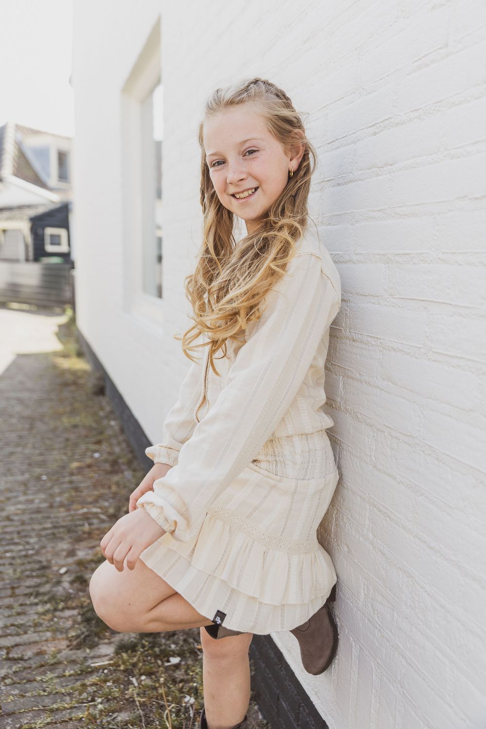 Portrait of a young person in a cream-colored ruffle dress leaning against a white wall on a paved walkway.