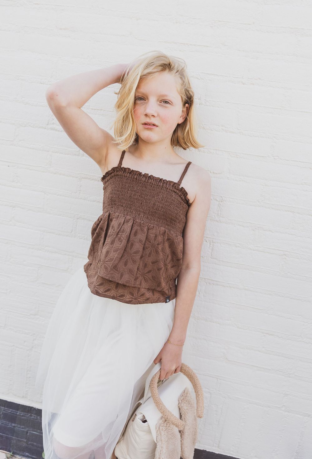 Young person wearing a brown smocked tank top and white skirt stands against white wall holding a teddy bear. Model fotografie