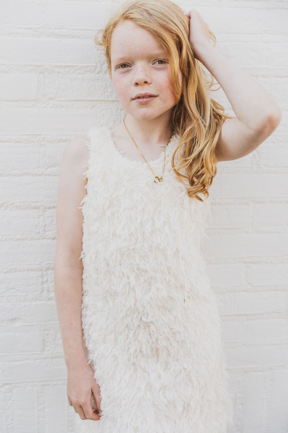 Young girl in white feathered dress poses against white brick wall while touching her strawberry blonde hair. kindermodel fotografie