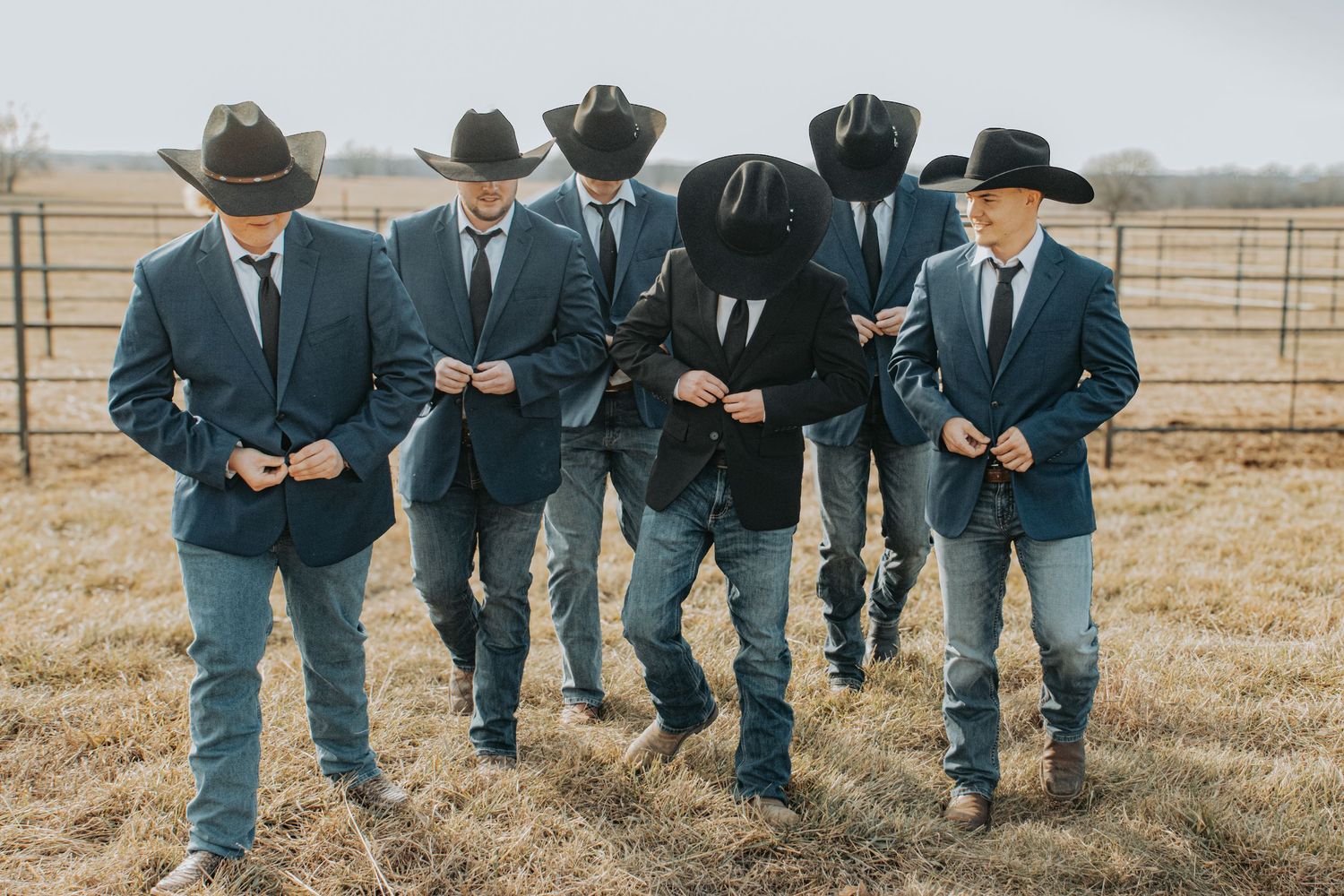 A group of people in matching navy blazers, cowboy hats and jeans walking together in a rural field setting.