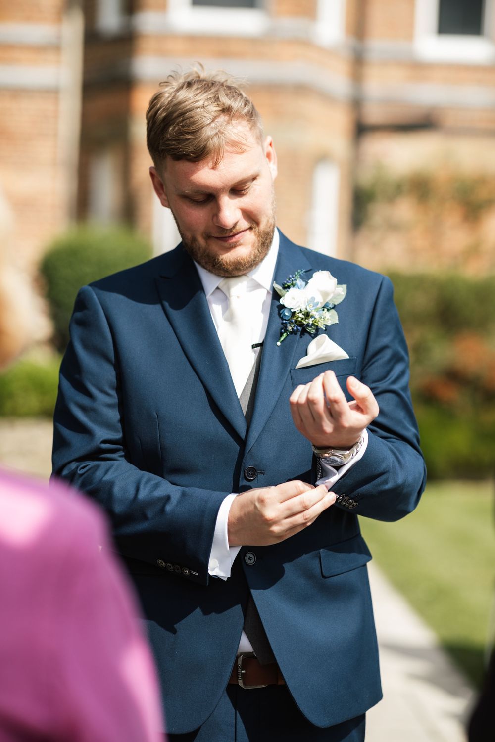 A formal wedding portrait of a groom in a navy suit adjusting his cuffs.