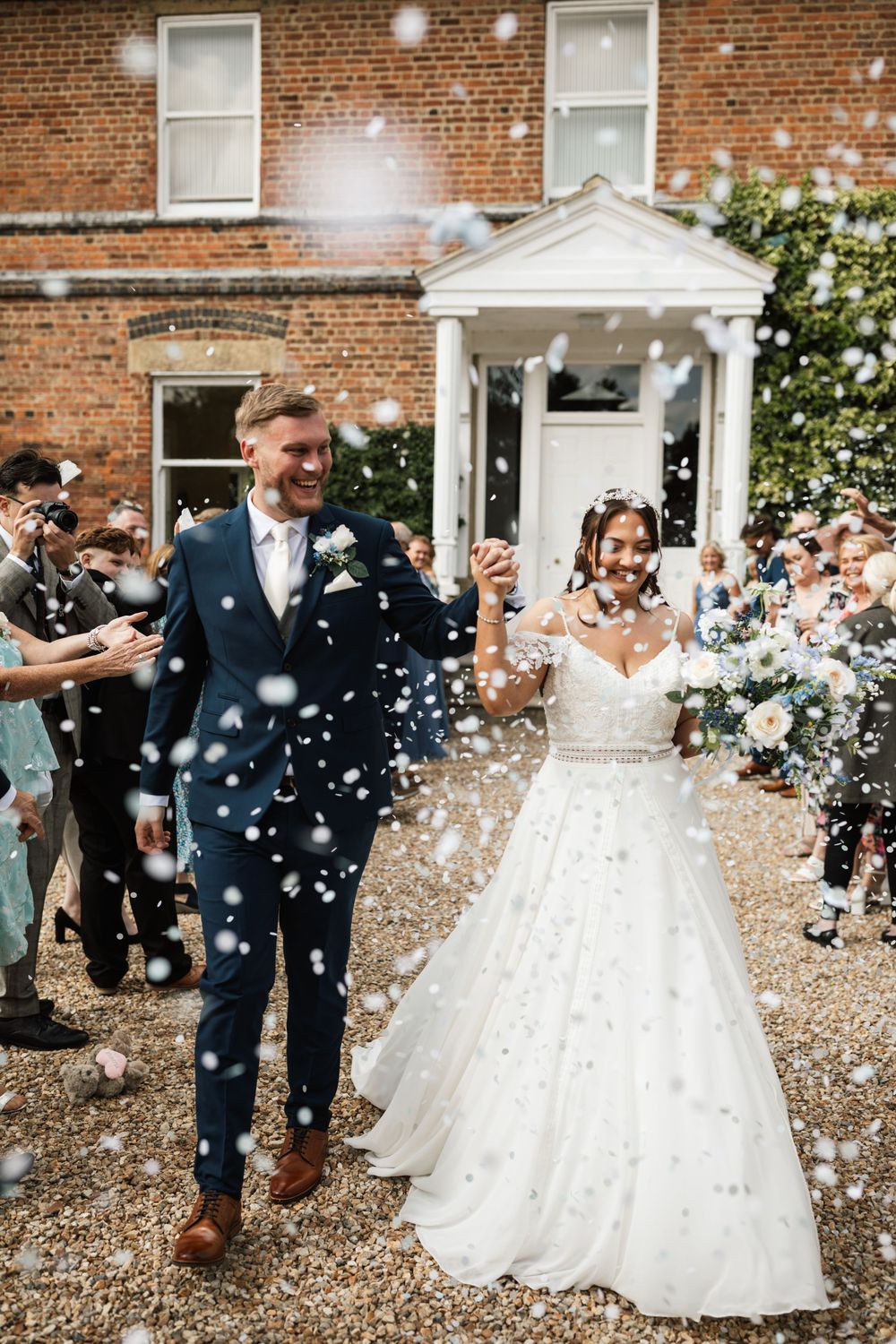 Newlyweds exit venue amid shower of white confetti against brick building backdrop.