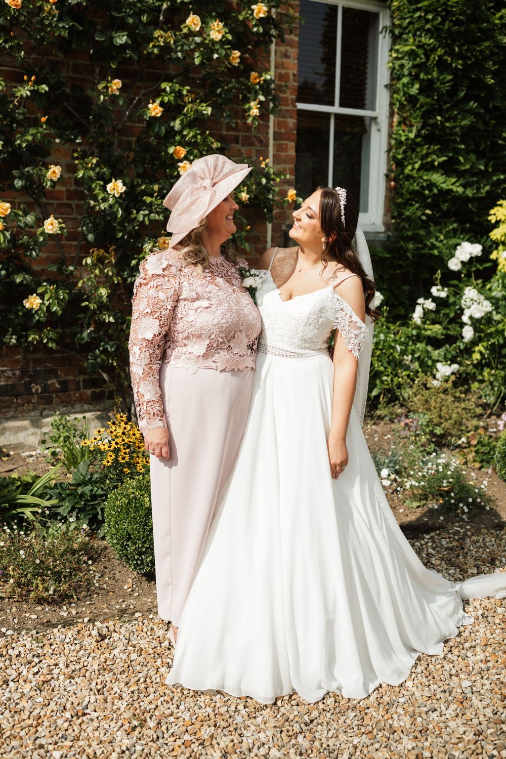 Bride and guest in pink dress share a moment among blooming garden roses.