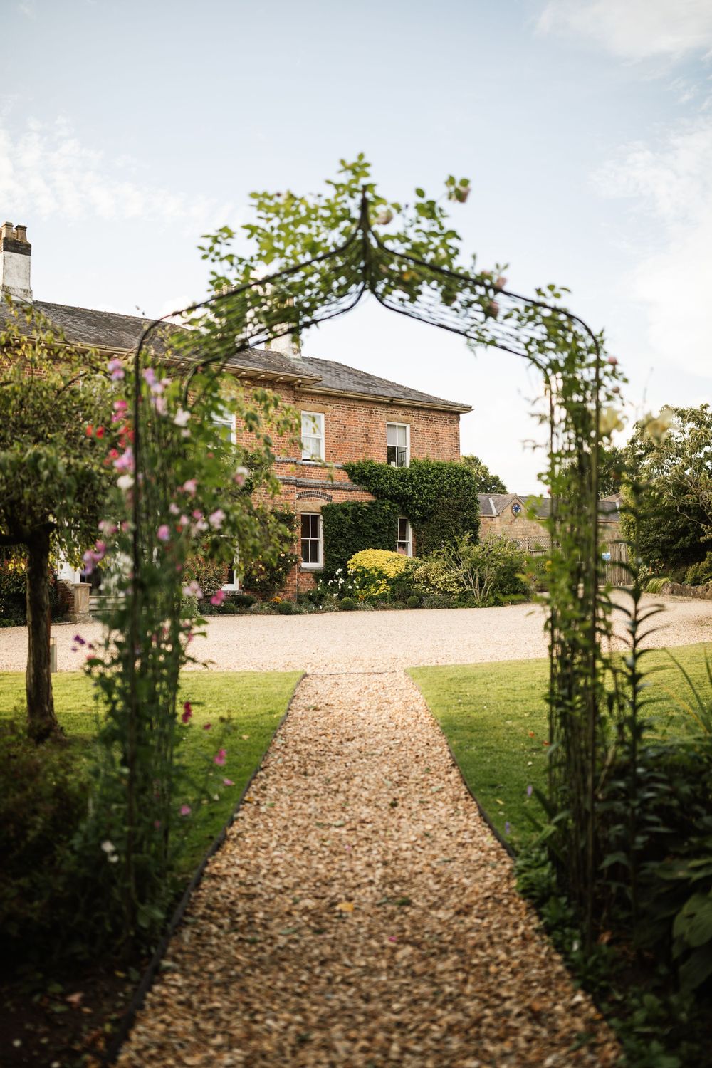 A stone pathway leads through a garden archway to a beautiful brick country house with manicured grounds.