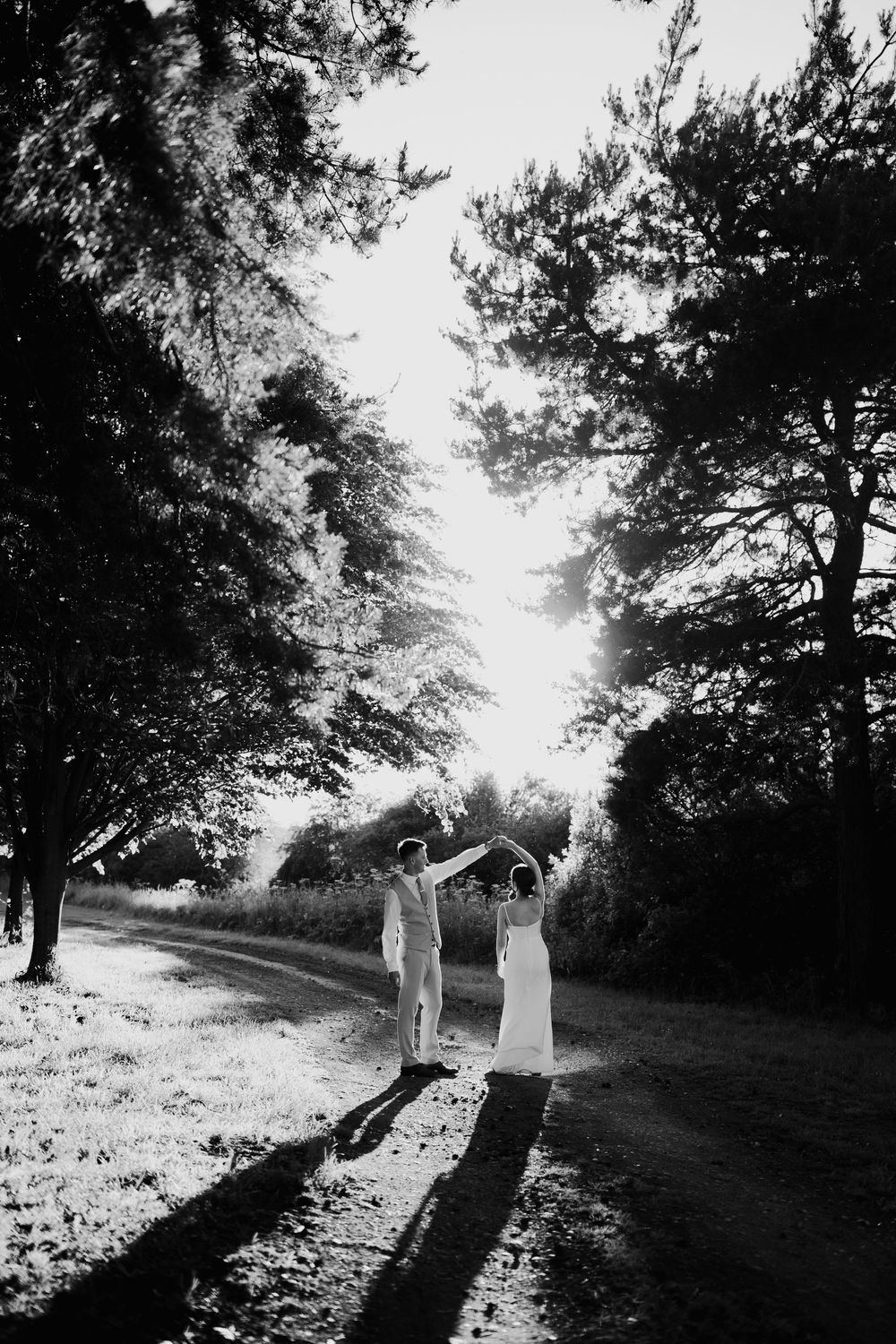 Black and white photo of newlyweds dancing on a tree-lined path in golden sunlight.