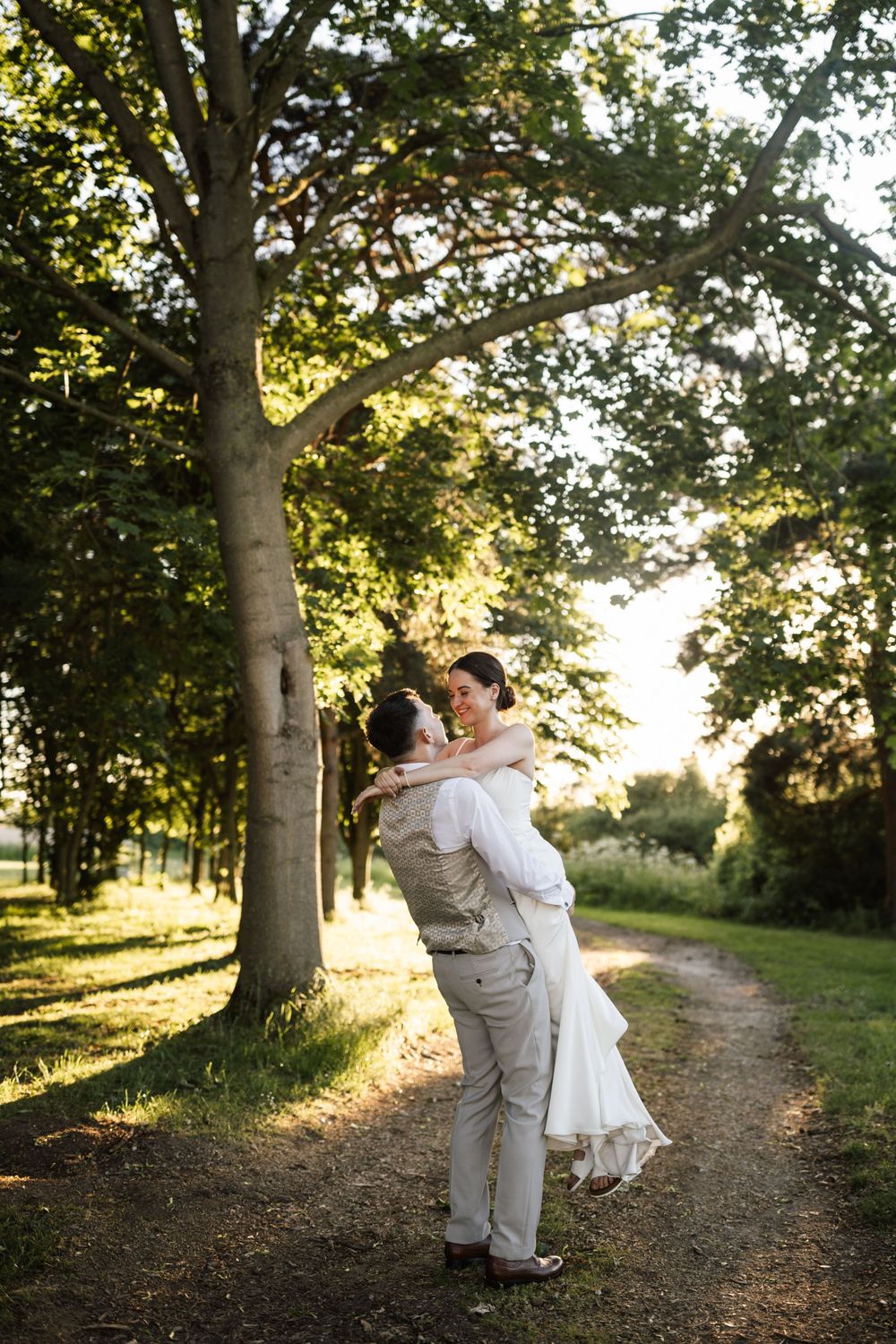 Groom lifting smiling bride on a sunlit garden path surrounded by lush green trees.