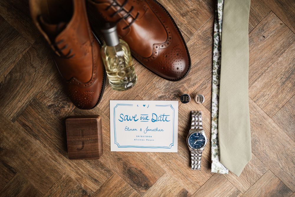Flat lay of groom wedding accessories including brown brogues, watch, tie, and save the date card.