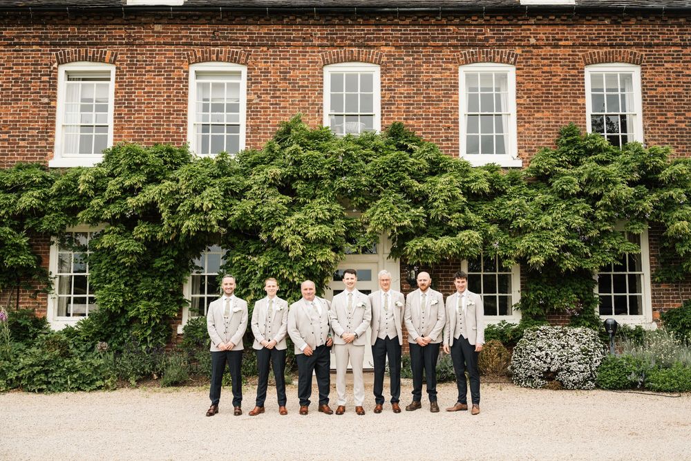 Groomsmen in beige suits stand together outside a brick manor house covered in ivy on a wedding day.