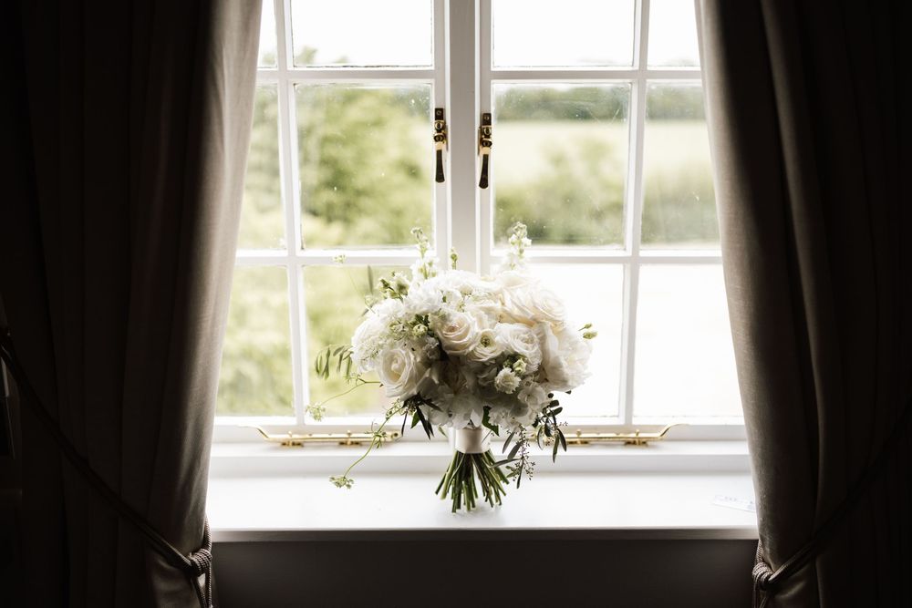 White bridal bouquet with roses and greenery hanging in front of a sunlit countryside window.