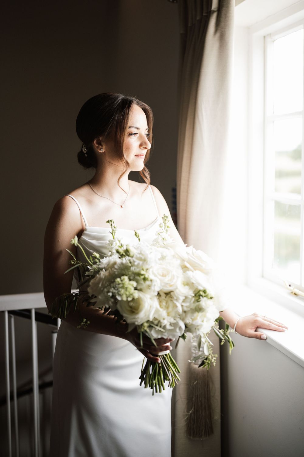 Bride in white satin dress holding lush white floral bouquet, gazing out sunlit window on wedding day.