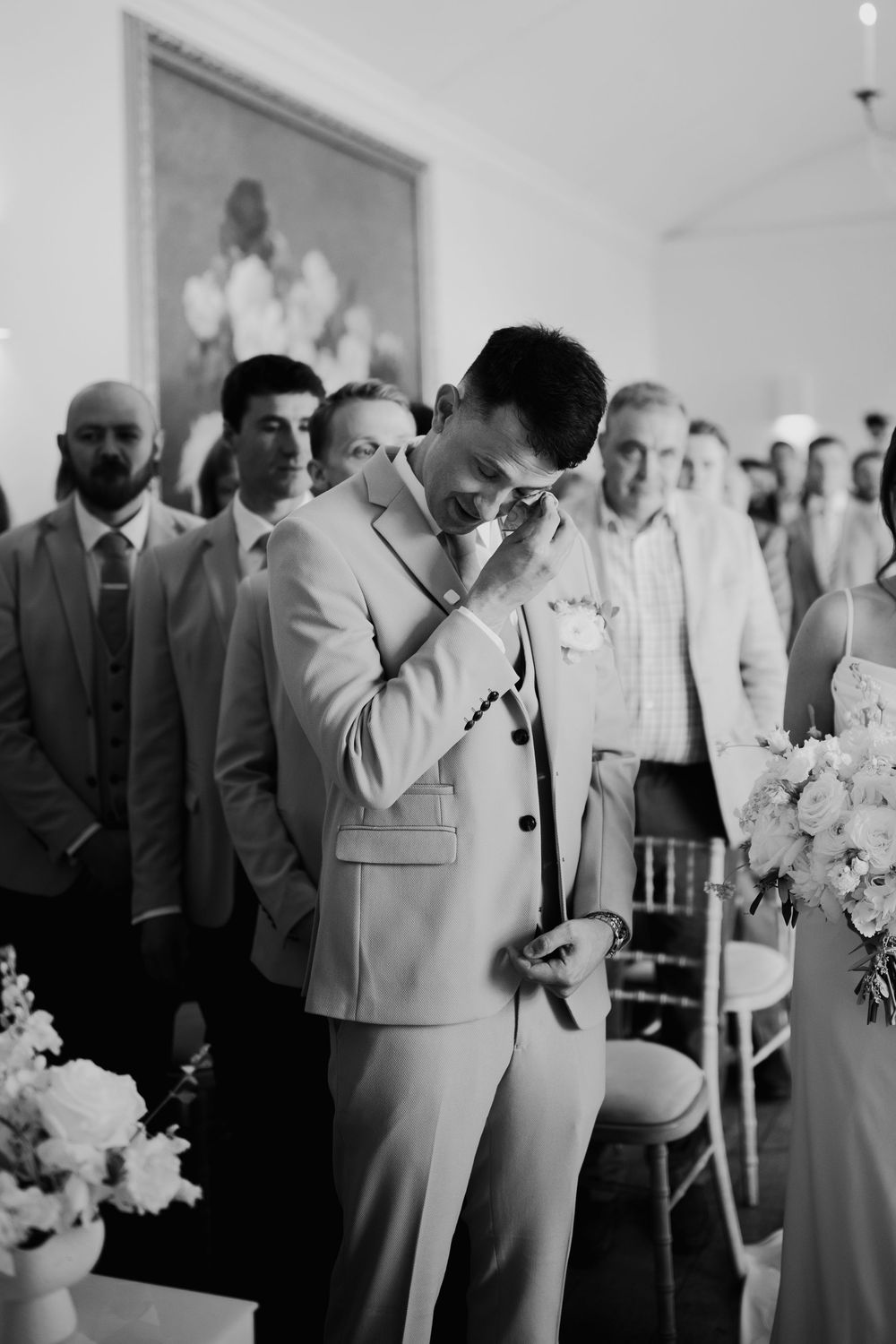 Emotional groom wiping tears as bride walks down aisle during wedding ceremony, black and white.