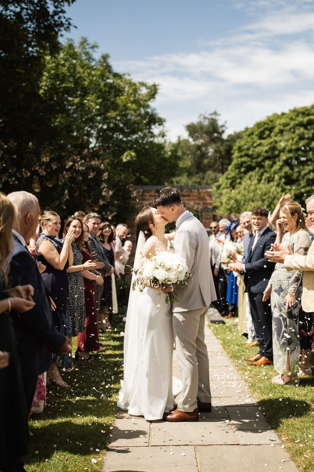 Bride and groom share a kiss while guests throw confetti outdoors on sunny wedding day.