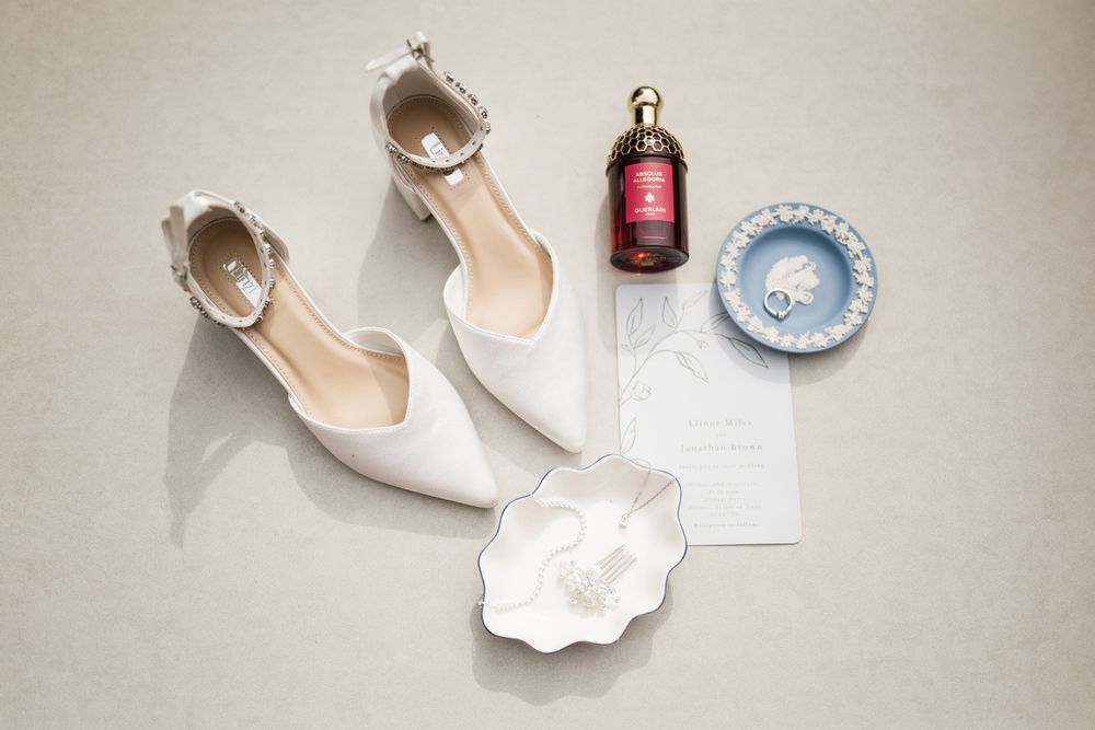 White bridal flats with ankle strap beside perfume bottle, blue dish, ring, and wedding invitation on beige surface.