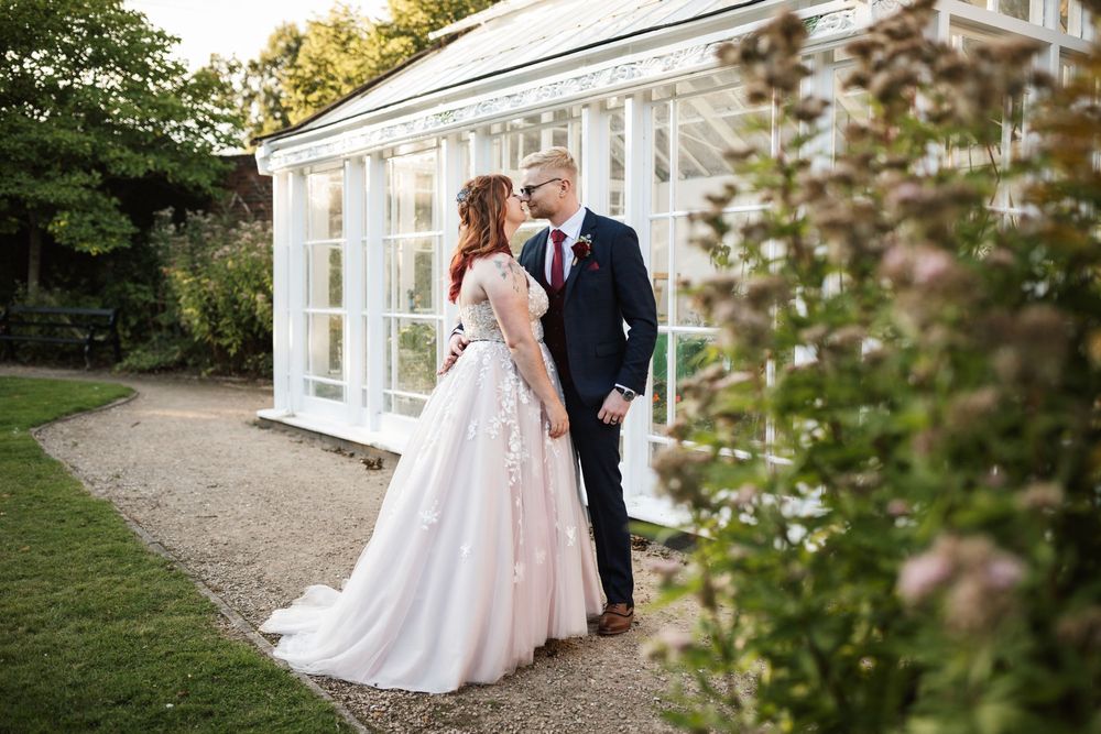 A couple shares a tender moment in front of an elegant white conservatory surrounded by blooming gardens at sunset.