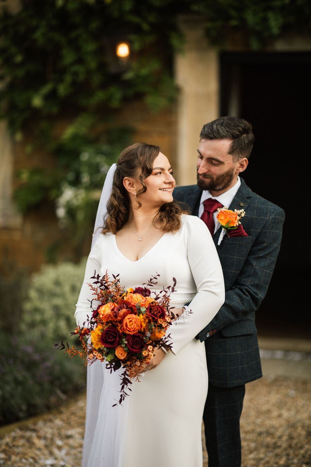 A couple in wedding attire standing close together outdoors with a vibrant orange and burgundy autumn bouquet.