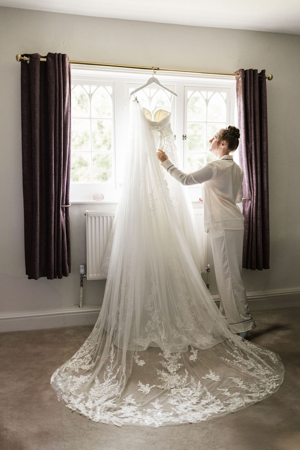 A wedding dress with lace train and cathedral veil is displayed in a room with dark curtains and arched windows.
