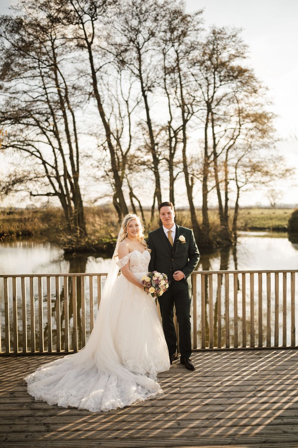 A newlywed couple stands on a wooden deck overlooking a serene lake surrounded by autumn trees at sunset.