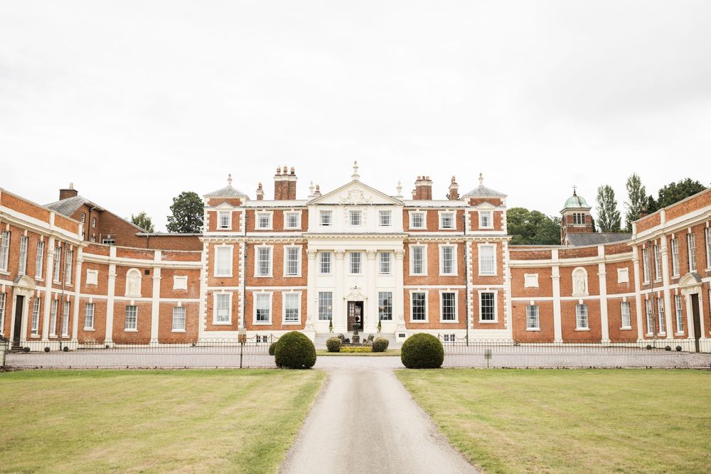 A grand Georgian mansion with symmetrical red brick wings and white center facade stands behind a manicured lawn with topiary.