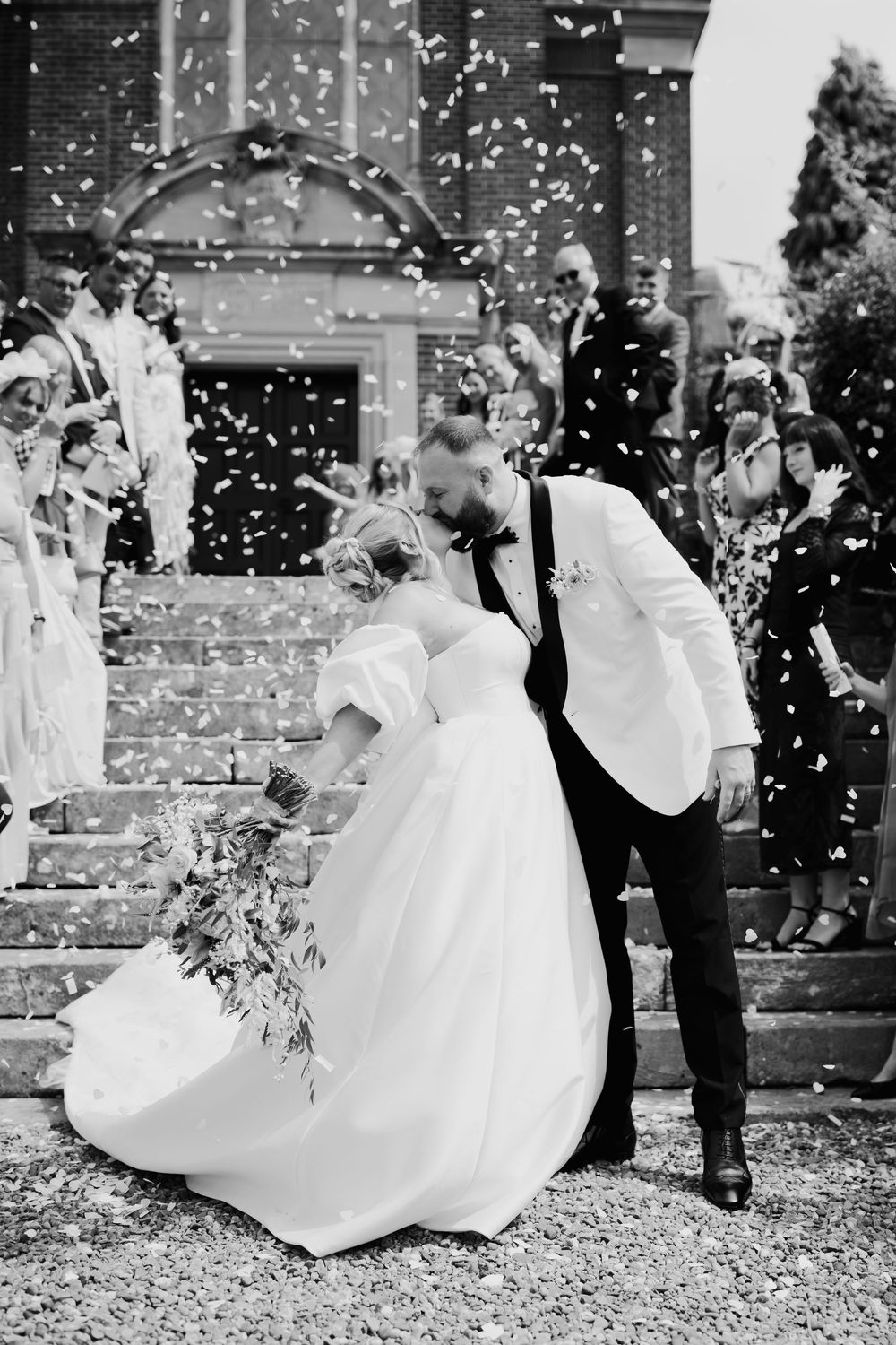 A black and white photograph of a romantic wedding moment with confetti falling on church steps in a joyful celebration.