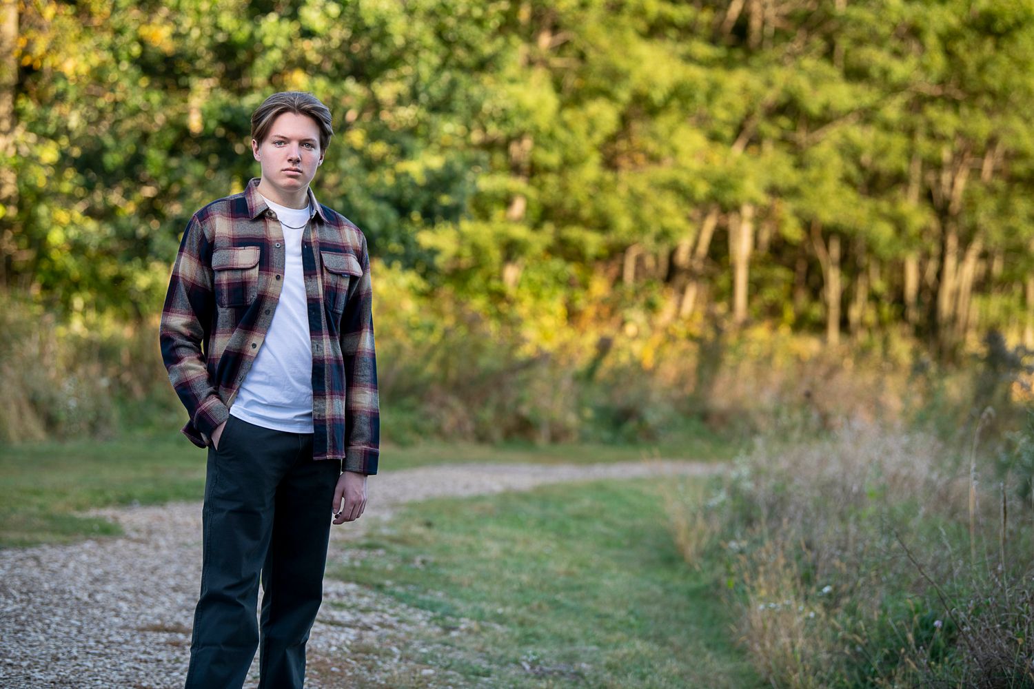 A person in a plaid flannel jacket and white t-shirt walks along a scenic dirt path surrounded by autumn foliage.