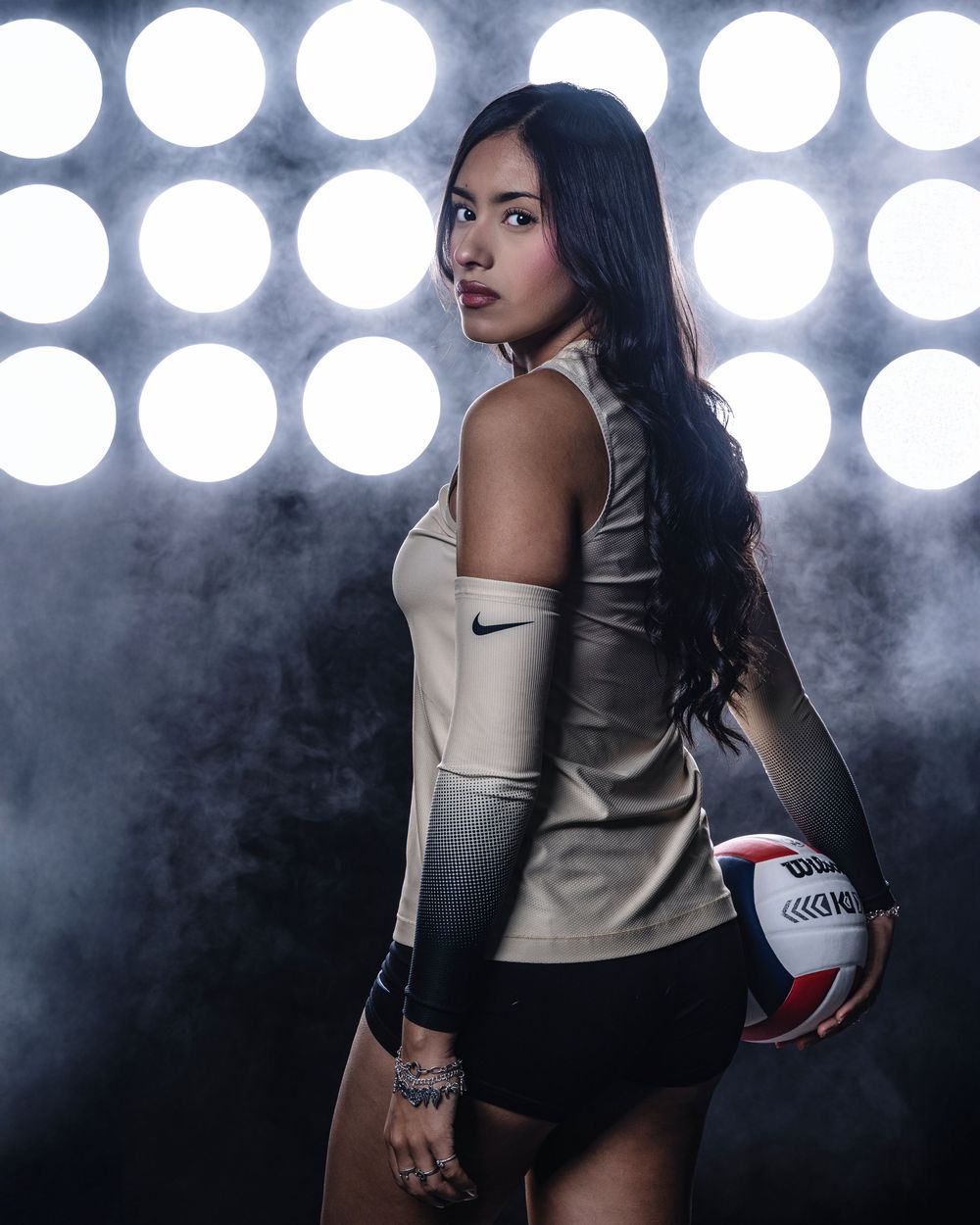 Athletic woman in black sports attire and metallic gold top poses dynamically with volleyball against illuminated backdrop.