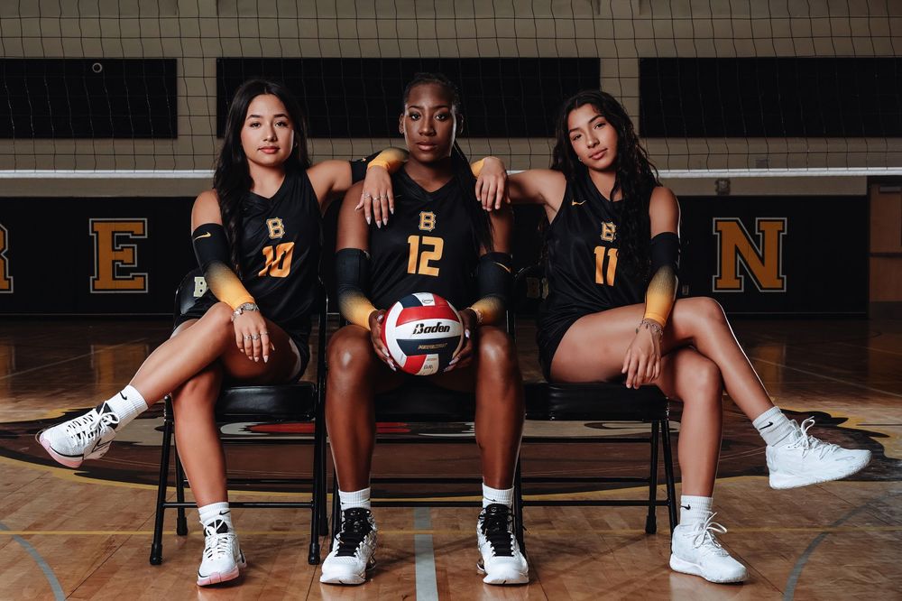 High school volleyball team players in matching black jerseys pose together on hardwood court with volleyball.