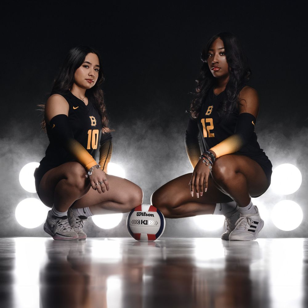 Two volleyball players in matching gold jerseys pose with a volleyball on a dark court with dramatic lighting.