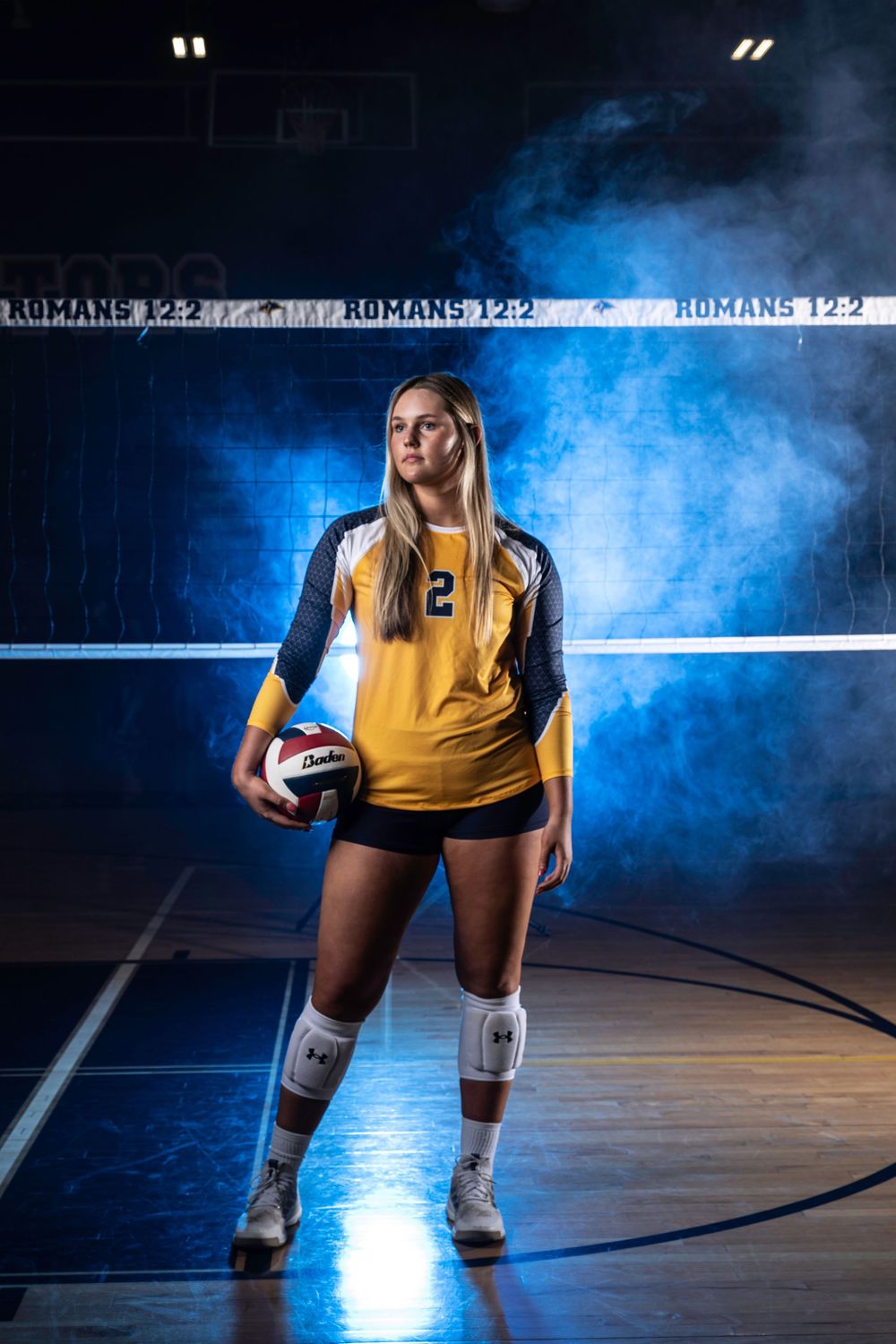 Volleyball athlete poses in yellow jersey against dramatic blue smoke on indoor court with volleyball.