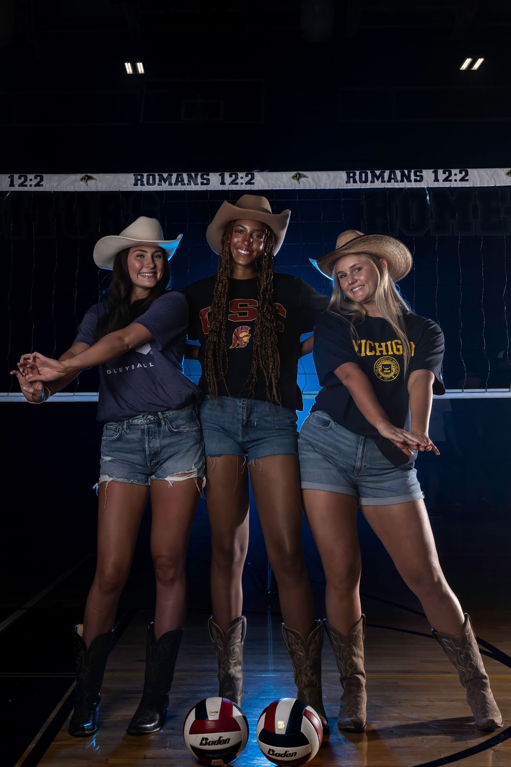 Three volleyball players in cowboy hats and denim shorts pose together on a court with volleyballs at their feet.