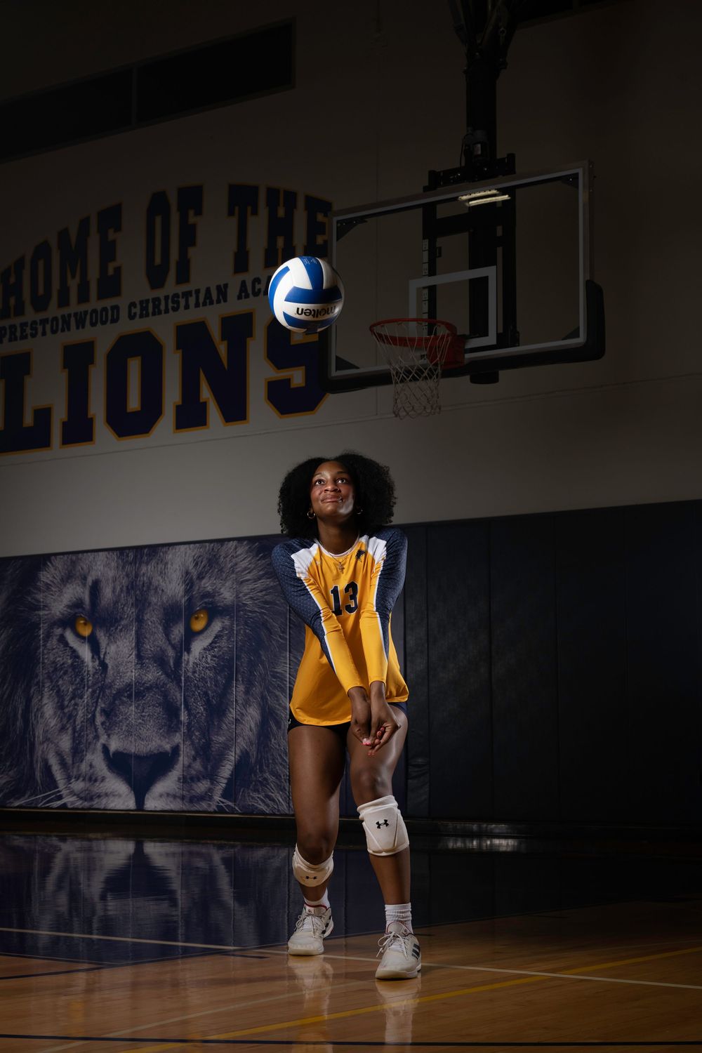 Athlete in yellow jersey stands on basketball court with volleyball and blue lion mural in background.
