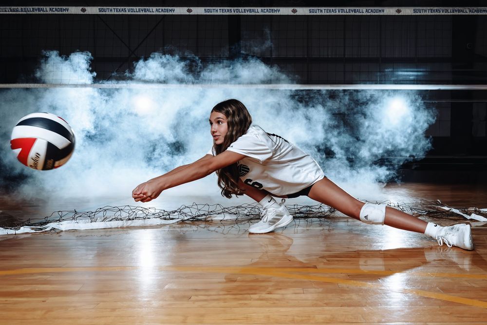 Indoor volleyball player diving to save the ball on hardwood court with dramatic smoke effect in background.