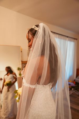 A bride in a white lace wedding dress and flowing veil stands in a sunlit room preparing for her ceremony.